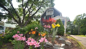 A house with a porch, surrounded by a garden with pink and yellow flowers. - Cottage 7 - Year Round - Pet Friendly $50 Fee - Stand Alone 1 Bedroom / 1 Bath (Wolfeboro)