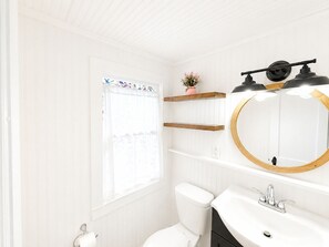 Een badkamer met toilet, wastafel en spiegel. Boven het toilet zijn houten planken en er is een raam met gordijnen.