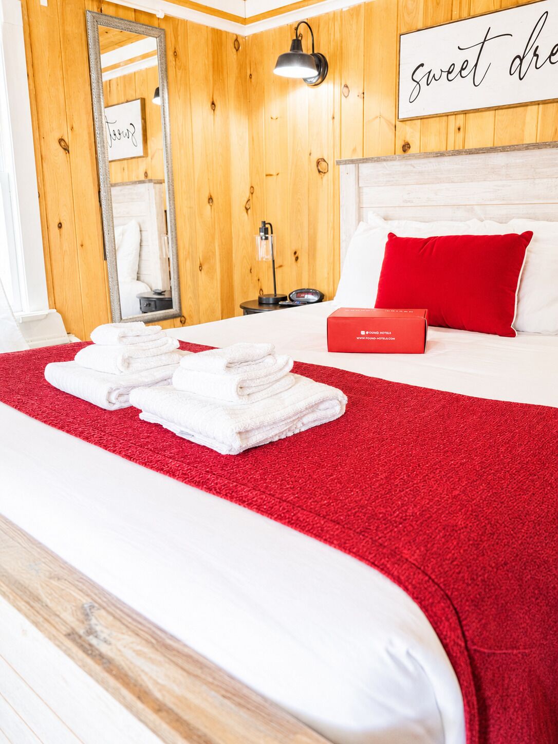 A hotel room with a bed, a red blanket, a red pillow, a bedside table with a lamp, and a sign on the wall.