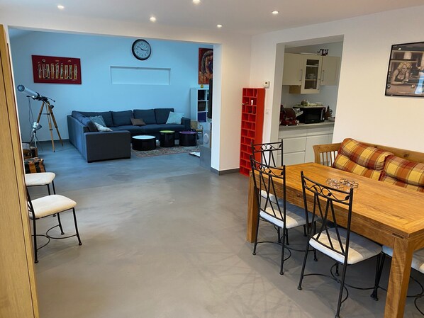 Uma sala de estar moderna com sofá, mesa de centro e mesa de jantar com cadeiras. Há uma área de cozinha com armários e uma estante vermelha.
