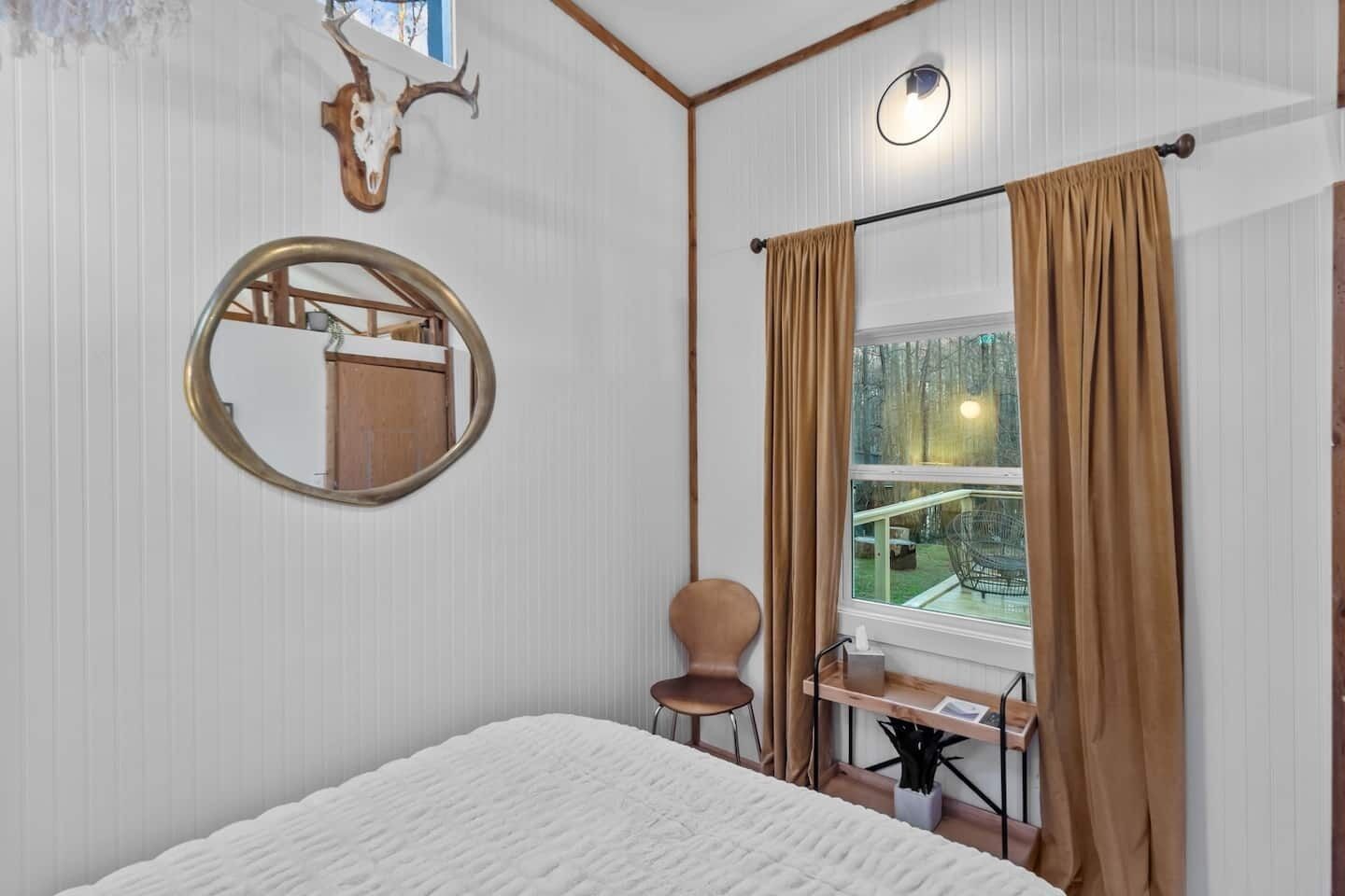 A bedroom with a bed, a round mirror, a wooden desk, a chair, and a window with curtains.