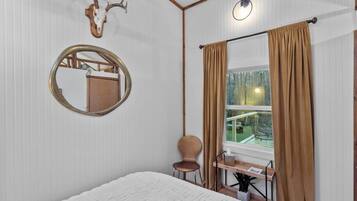 A bedroom with a bed, a round mirror, a wooden desk, a chair, and a window with curtains.