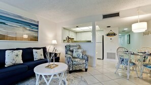 A living room with a dark blue sofa, a round white coffee table, and a patterned armchair. There is a dining area with white chairs and a glass table. The room has white walls and a ceiling with recessed lighting. - Coconuts 116 - Anna Maria Island - Condo -1 Bedroom/ 1 Bath- maximum occupancy of 6 people.     Distance to the Beach: 3 min walk  Community pool. (Holmes Beach)