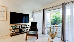 Una sala moderna con un televisor de pantalla plana, un sillón cómodo, una mesa de comedor y vistas a un jardín tropical a través de amplios ventanales.