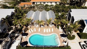 A resort with a swimming pool surrounded by lounge chairs and umbrellas, palm trees, and a building with a tiled roof.