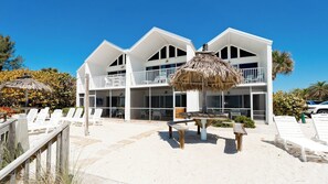 Un hotel frente a la playa de dos pisos con balcón, mobiliario exterior y una sombrilla de paja.