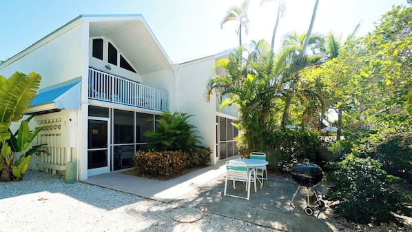 A modern two-story house with a balcony, a patio with a grill, and lush tropical vegetation.