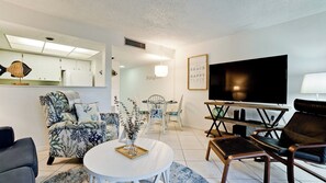 A modern living room with a flat-screen TV, a dining area with white chairs, and a sofa.