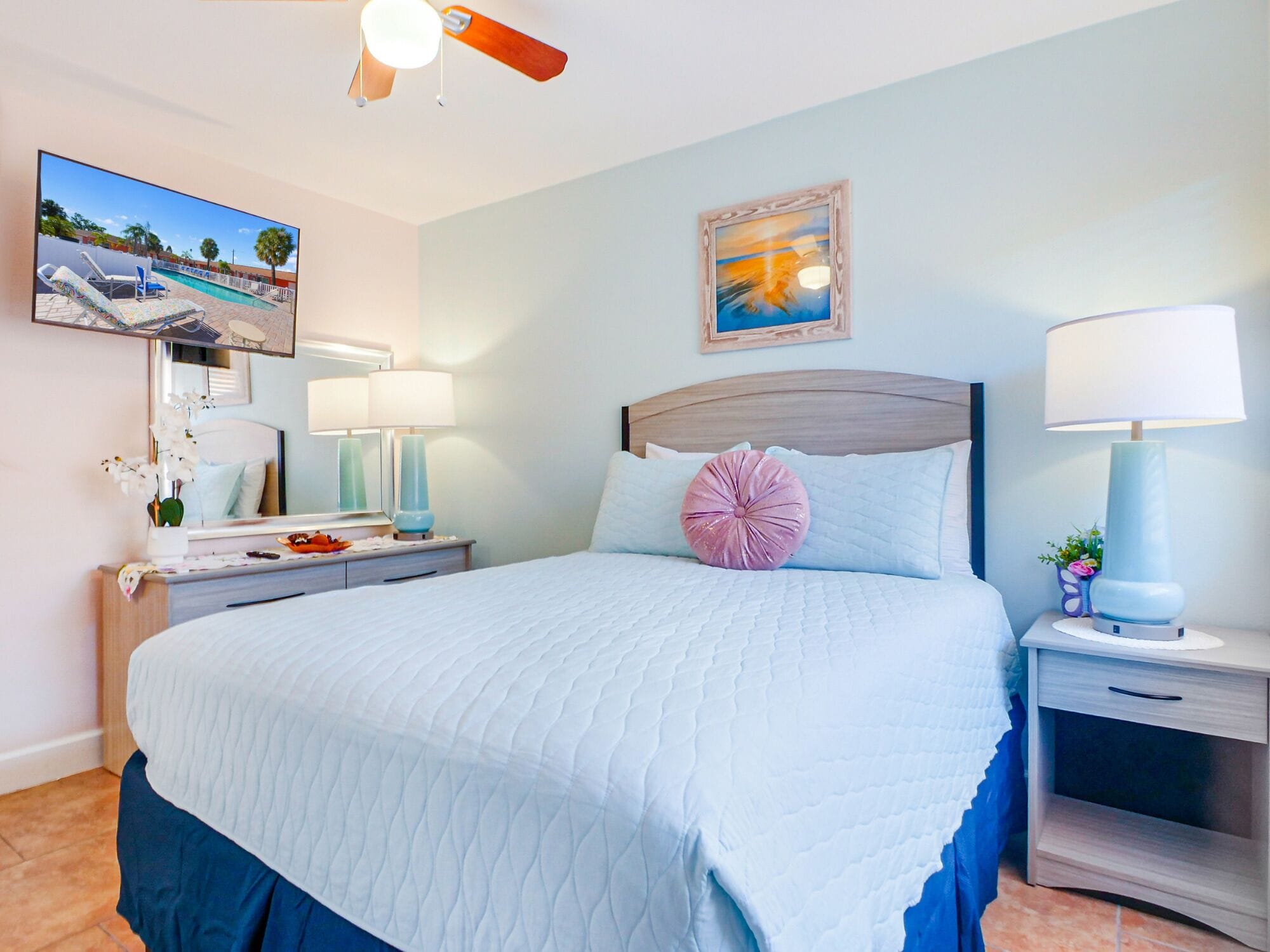 A bedroom with a bed, bedside tables, a nightstand, a ceiling fan, a painting, and a window with a view of a pool.