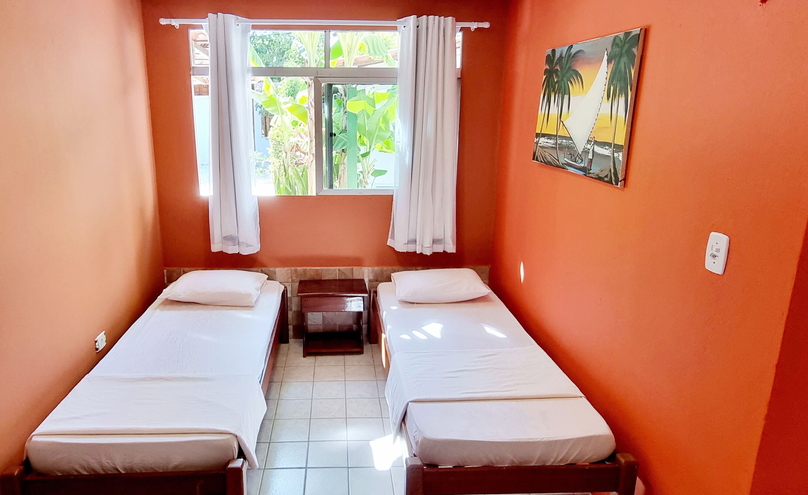Una habitación de hotel pequeña y con una cama individual, una ventana con cortinas, un cuadro en la pared y suelo de baldosas.