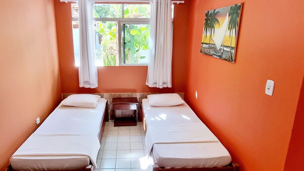A small, single-room hotel room with two single beds, a window with curtains, a painting on the wall, and a tiled floor. - Pousada Afrika (Ipojuca)