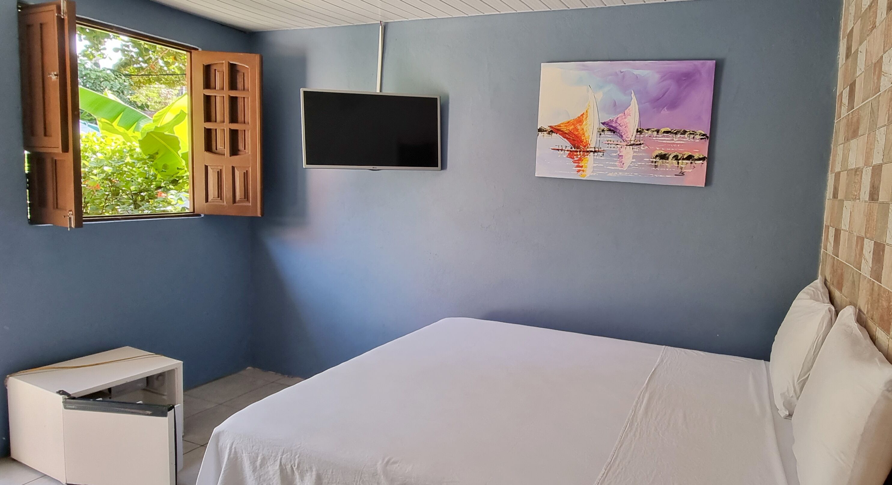 A bedroom with a bed, a TV mounted on the wall, and a window with open shutters.