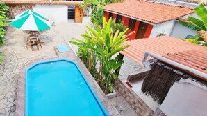 A swimming pool surrounded by stone tiles, a wooden table with chairs, and a red-roofed building. - Pousada Afrika (Ipojuca)