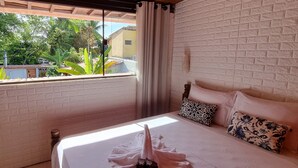 A bedroom with a bed, a window with curtains, a brick wall, and a view of greenery outside. - Pousada Afrika (Ipojuca)