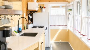Une cuisine compacte équipée d’un évier, d’une cuisinière et d’un four à micro-ondes. Une fenêtre avec des rideaux et une porte donnant sur l’extérieur sont également présentes.