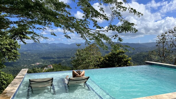 Una piscina cristallina con due lettini, circondata da alberi e un paesaggio montuoso.