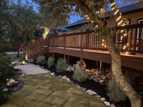A wooden deck with string lights, a small garden with rocks and shrubs, and a house in the background.