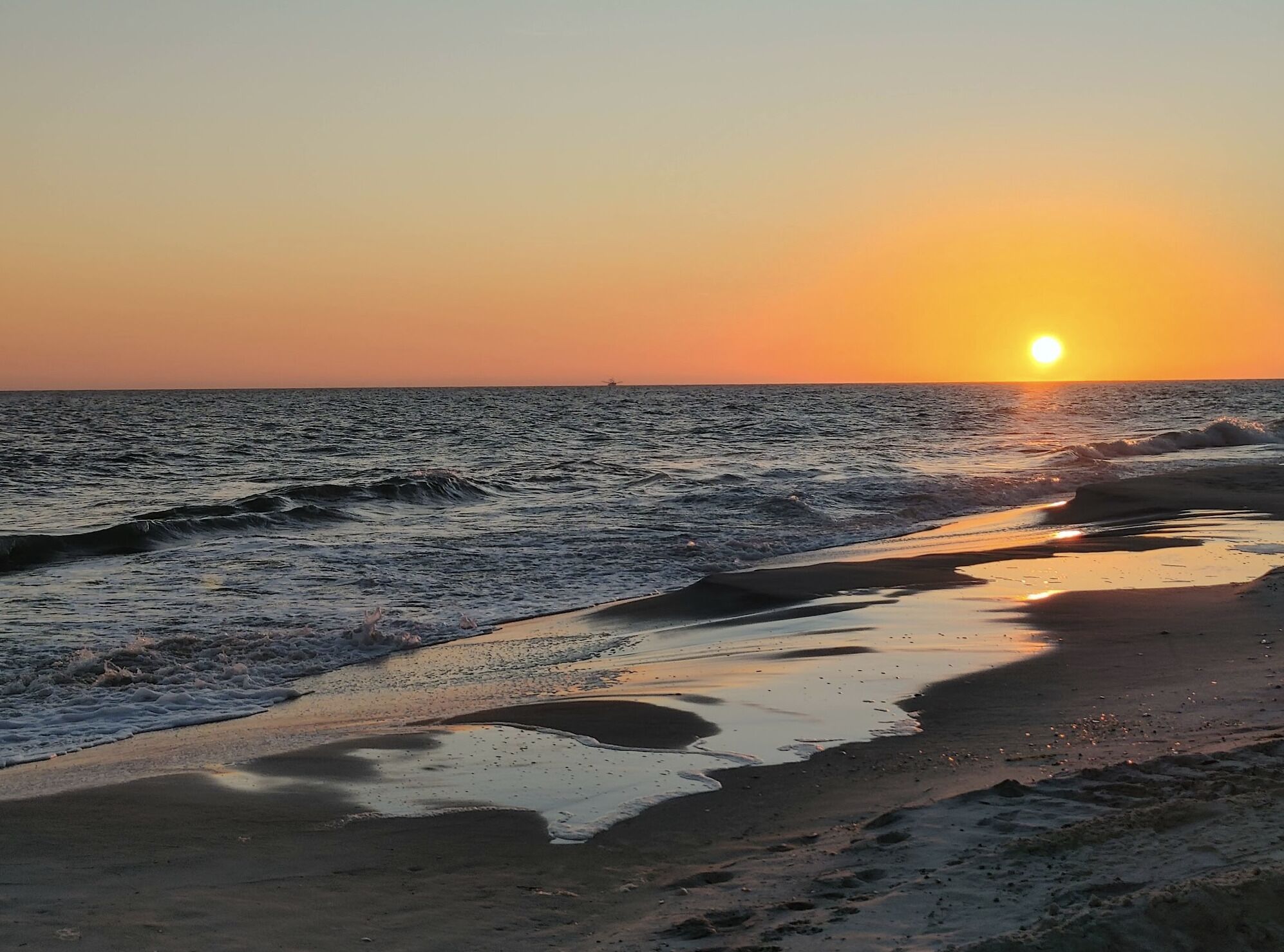 Une plage avec des vagues au coucher du soleil, un ciel dégagé et un bateau au loin.