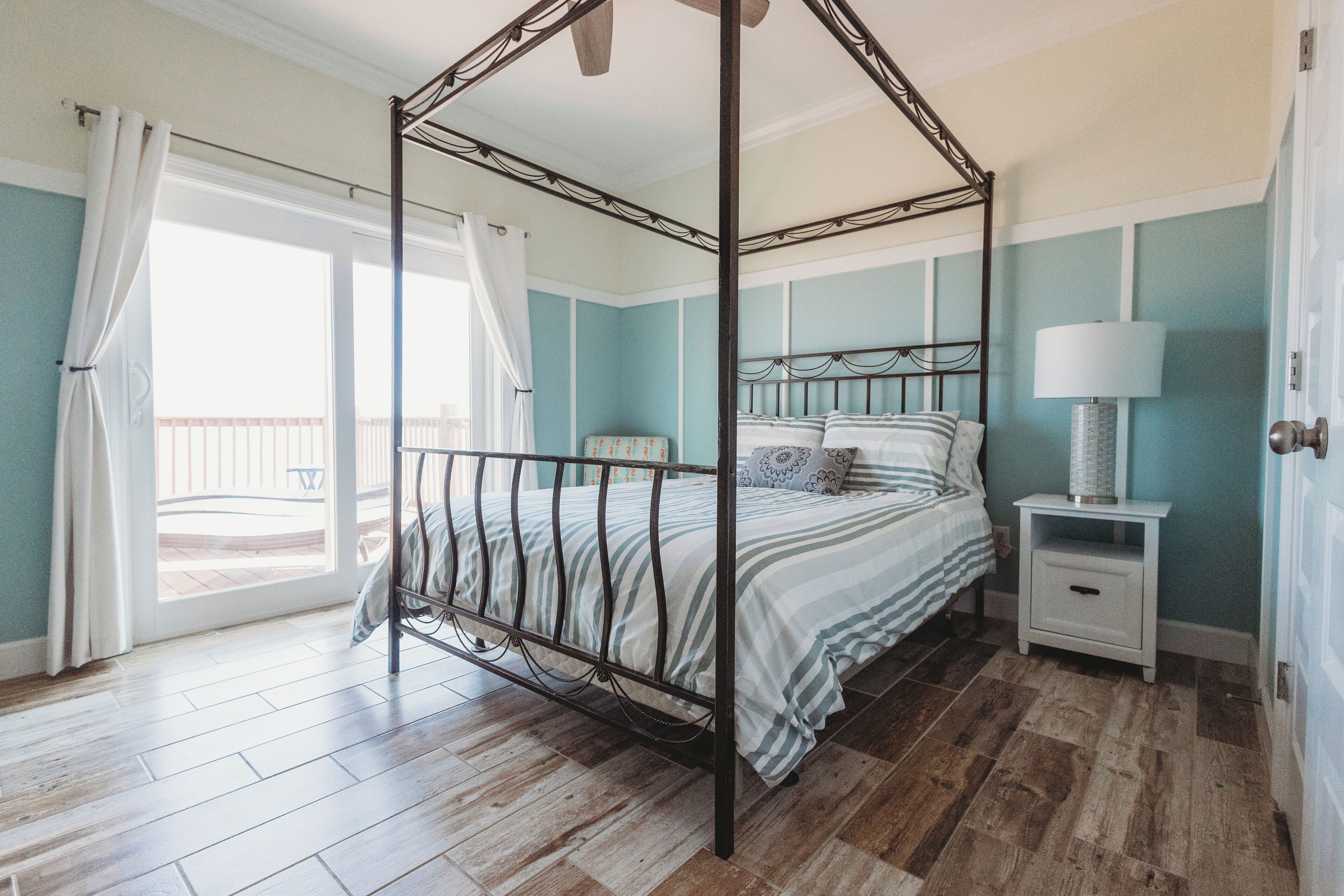 Un lit à baldaquin avec une couvre-lit rayée de blanc et de bleu, une table de chevet blanche avec une lampe, et une vue sur l’extérieur à travers une porte vitrée.