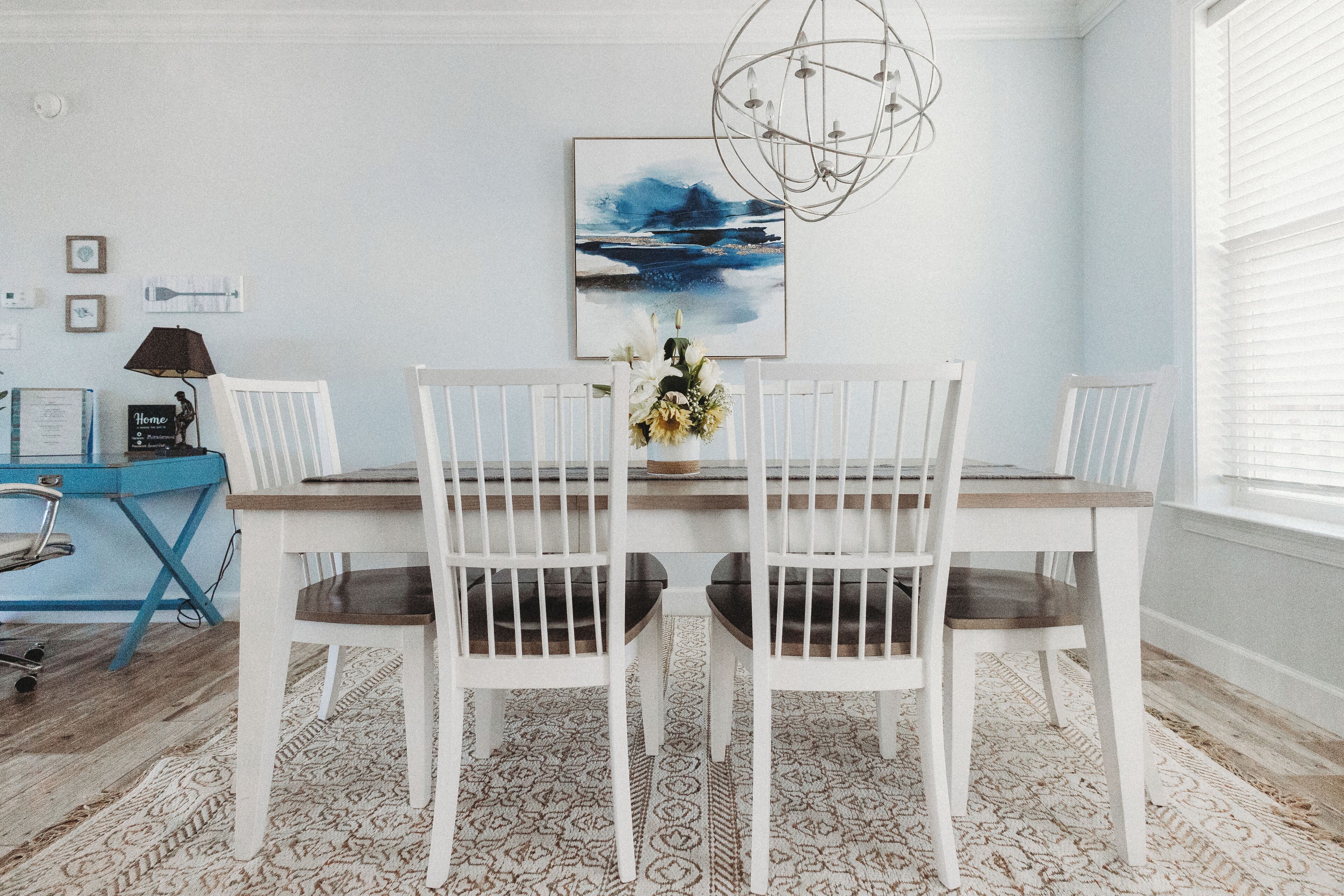 Un coin repas avec une table et des chaises blanches, un bureau bleu avec une lampe et un tableau accroché au mur.