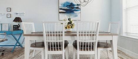 Un coin repas avec une table et des chaises blanches, un bureau bleu avec une lampe et un tableau accroché au mur.