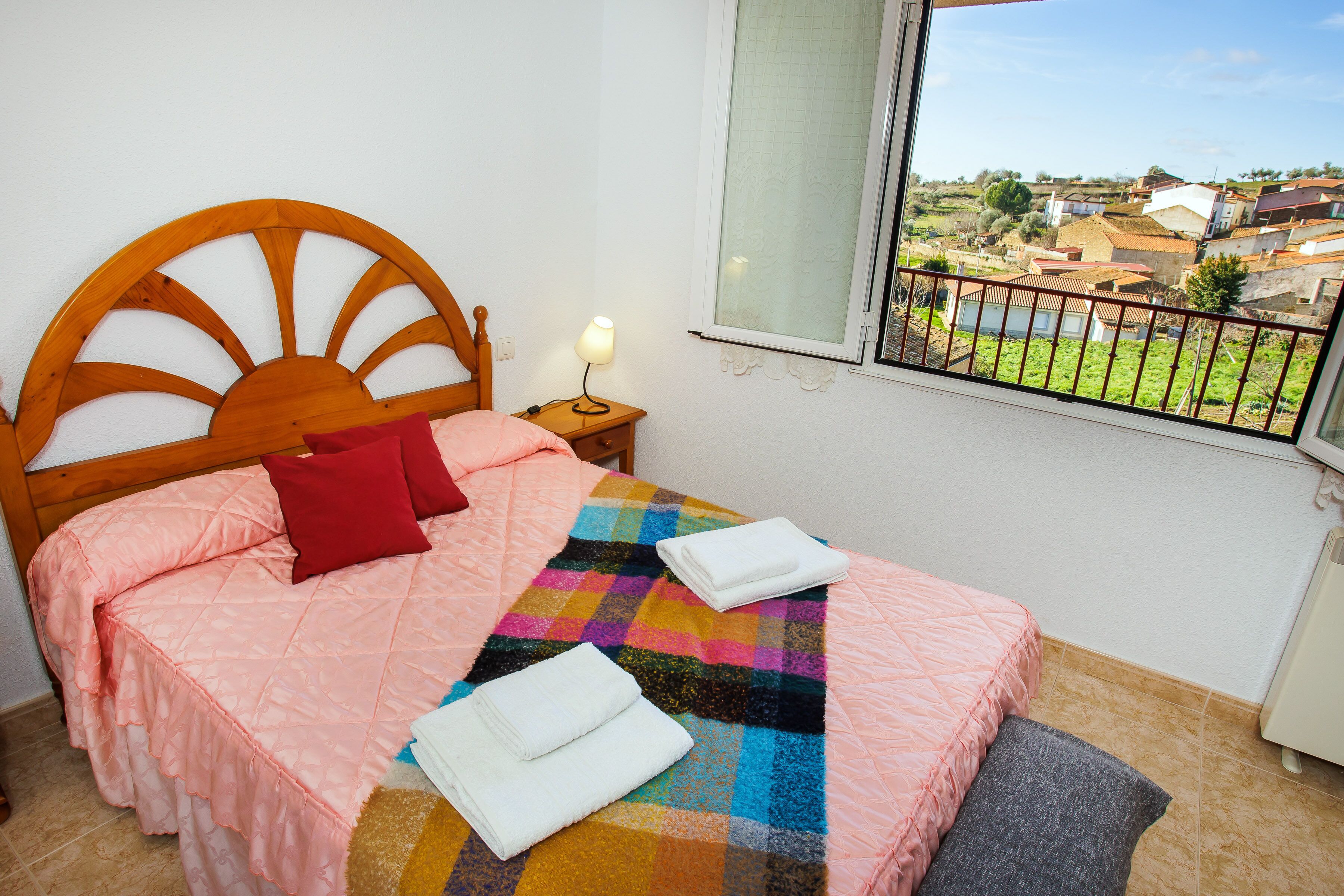 Une chambre à coucher avec un lit, une table de chevet, une fenêtre donnant sur un village et une balustrade de balcon.