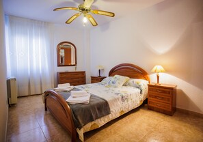 Une chambre à coucher avec un lit en bois, une table de chevet avec une lampe, un ventilateur de plafond, un miroir et une fenêtre avec des rideaux.