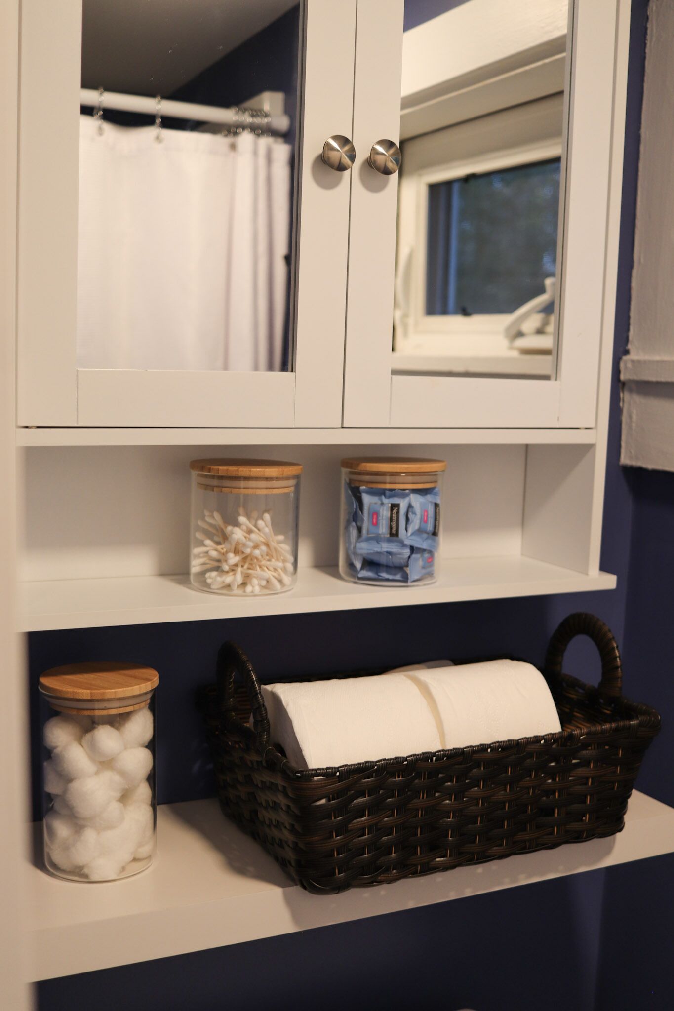 A bathroom with a white cabinet, a shower curtain, and two jars on the shelf.