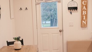 Interior acolhedor de uma cabana com teto de madeira, uma porta branca com janela, uma mesa de jantar com cadeiras e um letreiro com a mensagem 'Bem-vindo à Cabana'.