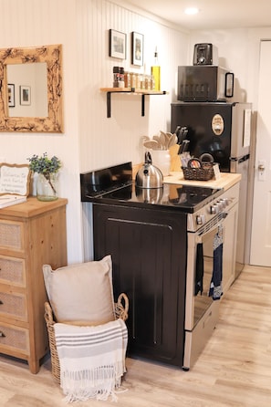 A compact kitchen with a stove, oven, and microwave. There is a wicker chair with a cushion and a small table with a sign. - NEW MaineStay Cottage #3 Full Kitchen + Fireplace in Hampden/Bangor (Hampden)