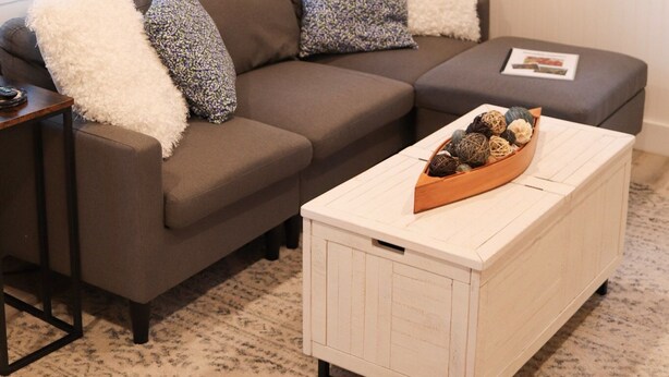 A living room with a brown sectional sofa, white throw pillows, a white coffee table with decorative items, and framed pictures on the wall.