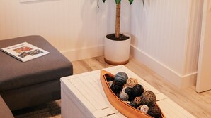 Uma sala de estar com televisão na parede, um vaso de planta, uma mesa em formato de barco de madeira com bolas decorativas, um sofá cinza e uma janela com persianas.
