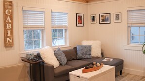 Uma sala de estar aconchegante com um sofá cinzento, almofadas brancas e fofas, uma mesinha lateral e uma mesa de centro branca com objetos decorativos. Há três quadros na parede e uma placa com a mensagem 'Bem-vindo à sua Cabana'.