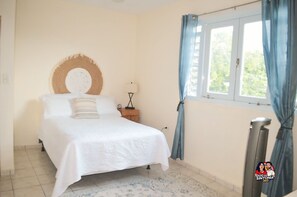A bedroom with a bed, a round wall decoration, a nightstand, a lamp, a window with curtains, and a radiator. - La Posada Mountain Retreat -  Exceptionally Stunning Views (Utuado)