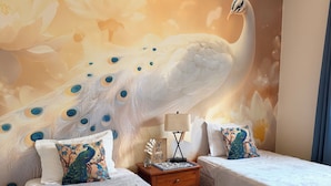 Une chambre avec deux lits simples, une table de chevet, une fresque murale représentant un paon et un ventilateur de plafond.