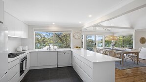 A modern kitchen with white cabinetry, a large island, and a dining area with a view. - Bush Retreat with Bayviews at Pretty Beach (HARDYS BAY)
