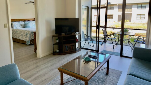 Uma sala de estar com sofá, mesa de centro de vidro, televisão sobre um móvel e varanda com mobiliário para área externa.