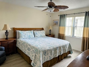 A bedroom with a bed, a wooden headboard, a nightstand, a lamp, a ceiling fan, and a window with curtains.