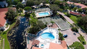 An aerial view of a residential area with a swimming pool, tennis courts, and a small dock by a water body.