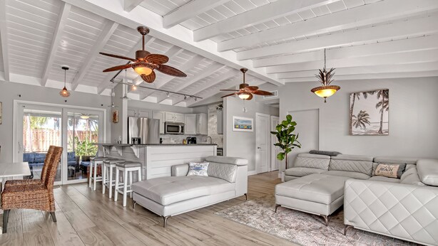 A spacious living room with a white sectional sofa, a dining area with a wicker chair, and a kitchen in the background. - Beach House just steps from Sun & Fun! (North Palm Beach)