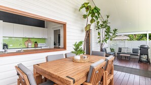 A modern kitchen with white cabinets and a green backsplash, a wooden dining table with chairs, and a patio area with a sofa and a grill. - High Three - Charming Beachside Retreat in the Heart of Yamba - Holiday Yamba (Yamba)