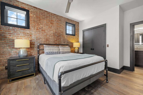 A bedroom with a brick wall, a bed with patterned pillows, bedside lamps, a wardrobe, and a door. - Nashville WeHo Escape | Walk to Bars & Dining (Nashville)