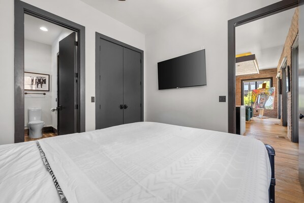A bedroom with a large bed, a flat-screen TV mounted on the wall, and a door leading to a bathroom.