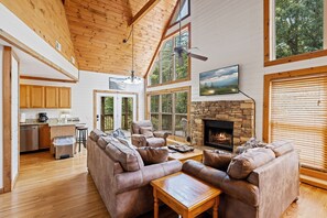 A spacious living room with a stone fireplace, wooden ceiling, and large windows.