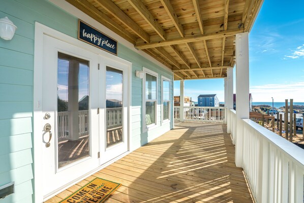 Une véranda couverte avec un plafond en bois et des balustrades blanches, une enseigne indiquant « endroit où le bonheur règne », et une vue sur la plage et l’océan en arrière-plan.