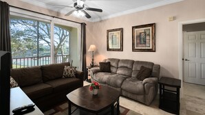 A living room with a brown sofa, a coffee table with flowers, a ceiling fan, and two framed pictures on the wall. - Spacious 3 bedroom, 2 bathroom condo in a great community! (Kissimmee)