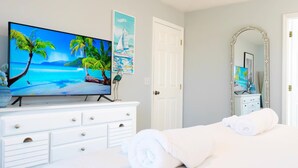 A bedroom with a TV displaying a tropical beach scene, a white dresser, a bed with rolled towels, and a large mirror.