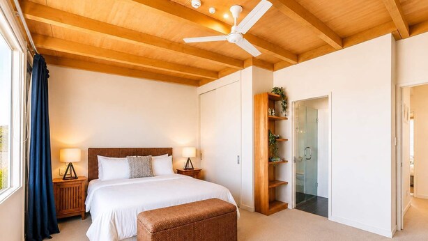 Un dormitorio con cama, lámparas de noche, ventilador de techo, un banco de madera y un vidrio que da al baño.