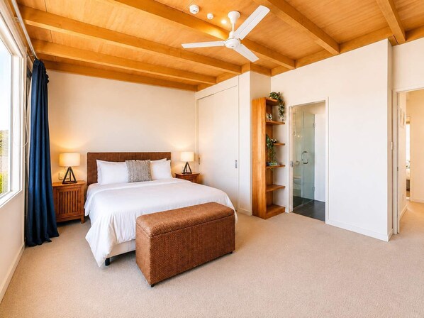 A bedroom with a bed, bedside lamps, a ceiling fan, a wooden bench, and a glass door leading to a bathroom.