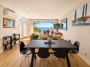 A modern living room with a dining area, large windows, and a view of the sea.